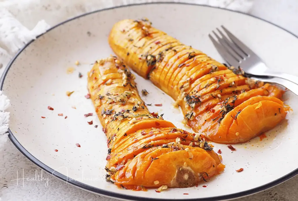 hasselback cut butternut squash on a white plate topped with herbs and seeds.
