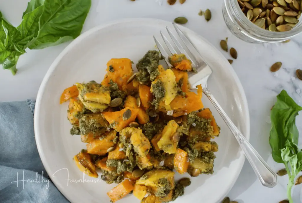 Round white plate with cubes of butternut squash covered in green pesto and pumpkin seeds.