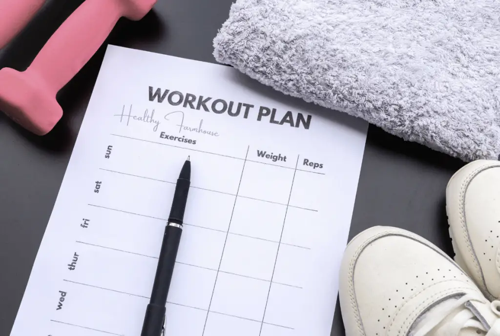 Pink hand weights, grey towel, white tennis shoes, and a blank workout plan sheet for working out throughout the day featuing a black pen laying on top.