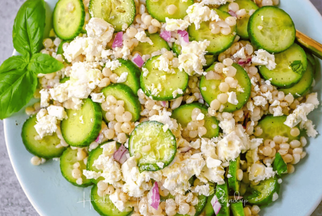 Bright green cucumber salad with large pear couscous and feta cheese sprinkles placed in a white dish with garnish